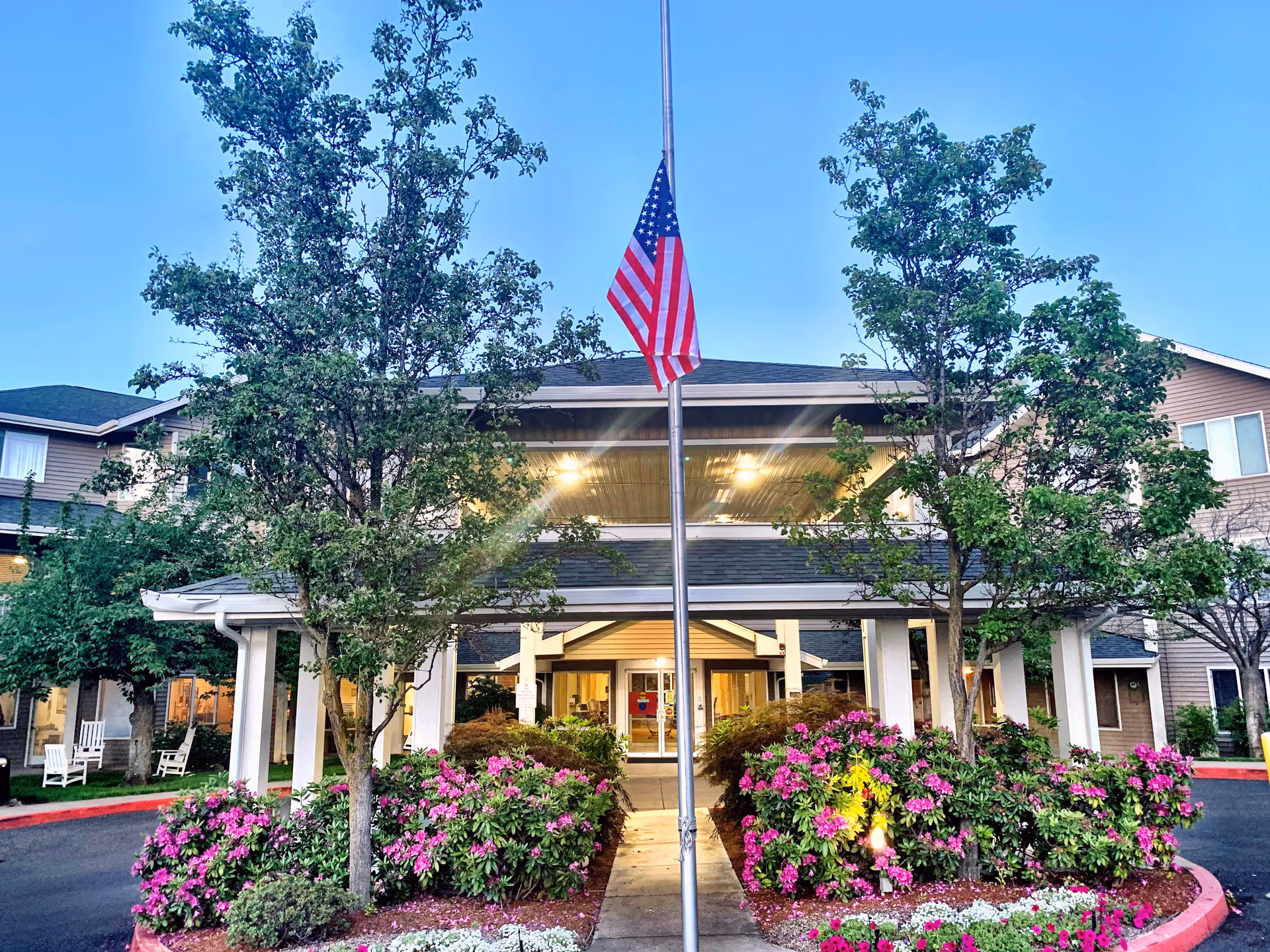 Front exterior view of Maple Ridge Senior Living facility with a covered entrance, an American flag on a flagpole at half-mast, trees, and colorful flower beds in the foreground.