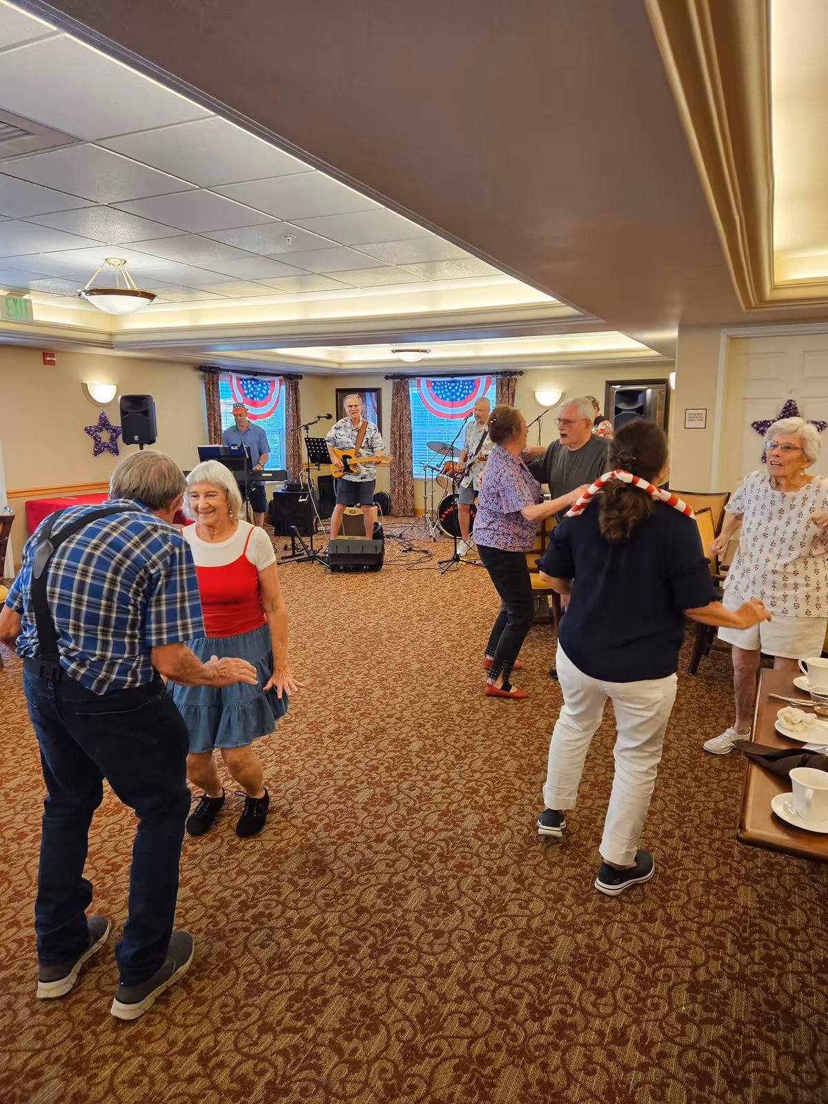 A group of elderly people dancing and socializing in a carpeted room with a live band playing music in the background. The room has patriotic decorations with red, white, and blue bunting on the windows. There are tables with cups and plates on the right side of the image.