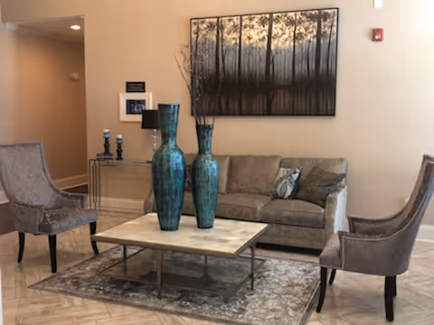 A cozy living room area with a beige sofa adorned with two decorative pillows, two upholstered chairs facing each other, a rectangular coffee table with two large blue vases, and a patterned rug underneath. A large framed artwork depicting a forest scene hangs on the wall behind the sofa. A console table with candles and a lamp is visible in the background.