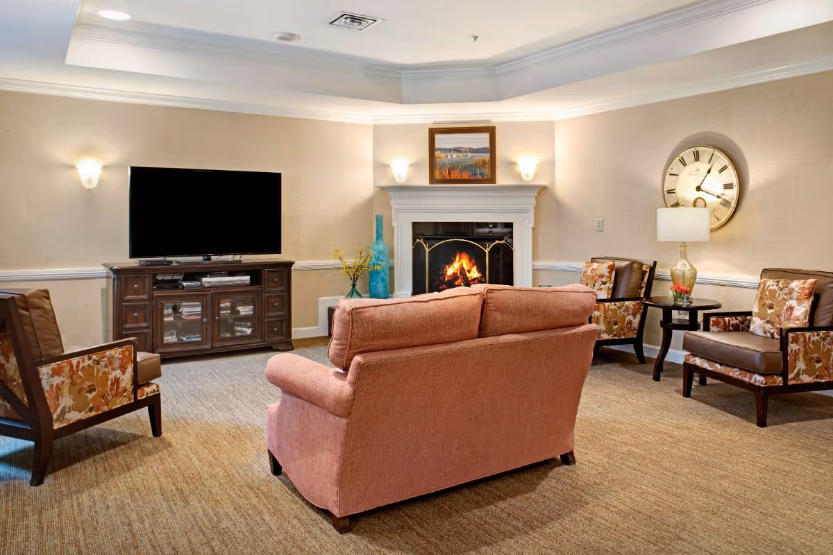 A cozy living room with a pink sofa facing a fireplace with a fire burning inside. There are two floral-patterned armchairs and a wooden TV stand with a flat-screen TV. The walls are beige with white trim, and there is a large clock and a table lamp on a side table.