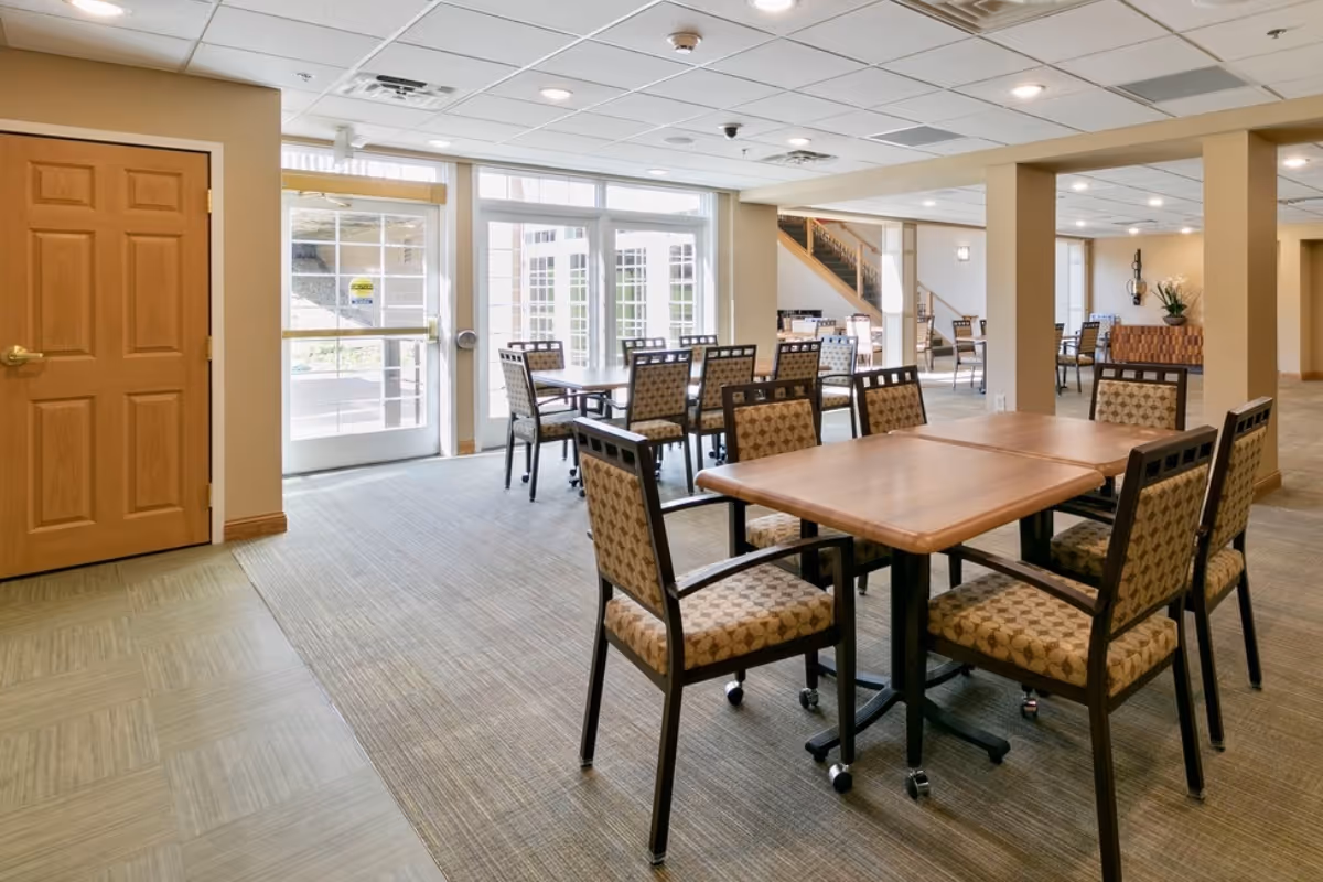 Bright communal dining area with wooden tables and patterned chairs by large windows and double doors.