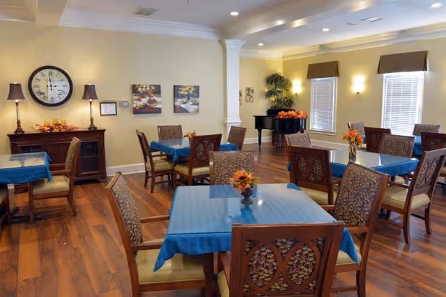 A dining room with several wooden tables covered with blue tablecloths and decorated with small flower arrangements. The room has wooden flooring, a sideboard with two lamps and a large clock on the wall, two framed pictures, a black grand piano in the corner, and windows with brown valances letting in natural light.