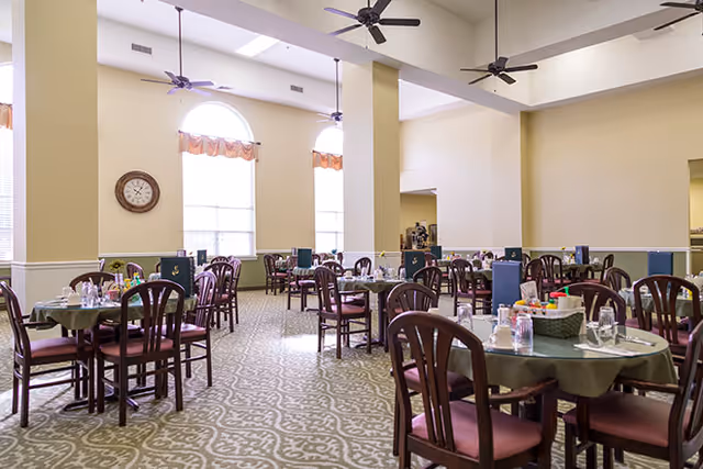 A spacious dining room with multiple round tables covered with green tablecloths, each set with glasses, plates, and utensils. The room has high ceilings with ceiling fans, large arched windows with curtains, and a wall clock. The floor has a patterned carpet, and the walls are painted in light colors.