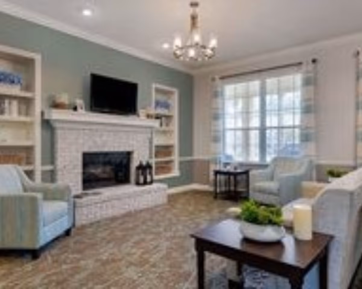 Bright furnished living room with a fireplace, wall-mounted TV, armchairs, coffee tables, and a large window.