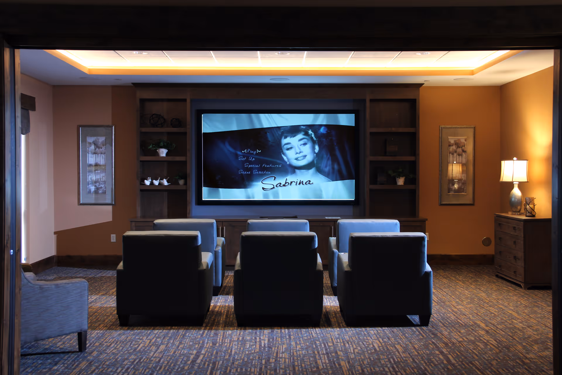 Cozy screening room with six upholstered chairs facing a large wall-mounted screen showing the movie Sabrina.