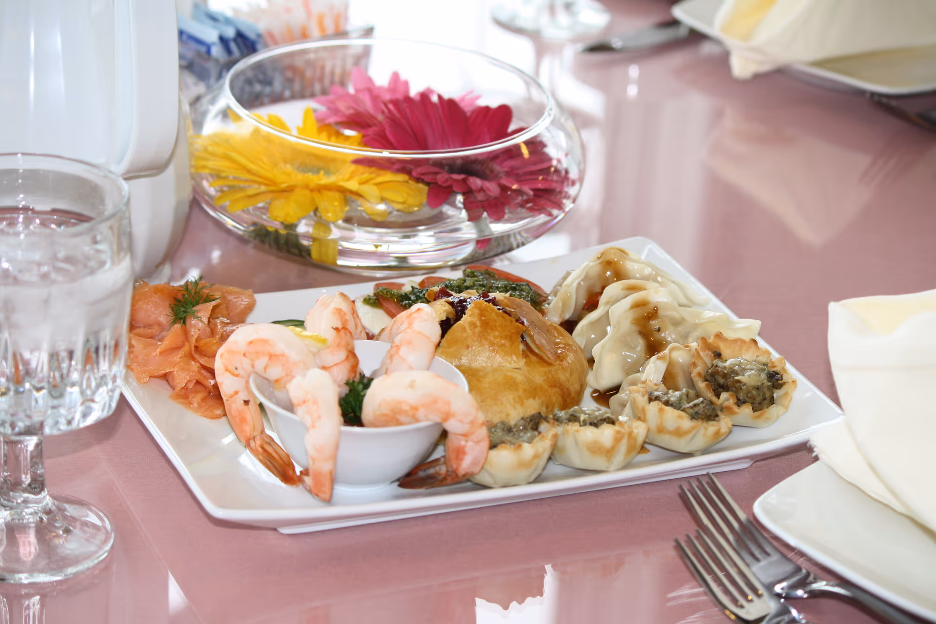A platter of shrimp, dumplings, and small pastries on a dining table with a glass of water and a floral centerpiece.