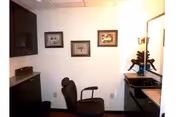 Small room with a black salon chair in front of a counter with a sink and a mirror. The walls are white with three framed pictures hanging. There are dark cabinets on the left side and a small box on the counter.