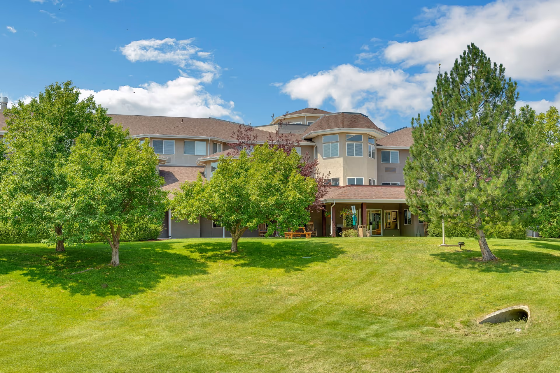 Exterior view of a multi-story senior living facility building with a sloped green lawn and several trees in the foreground under a partly cloudy blue sky.