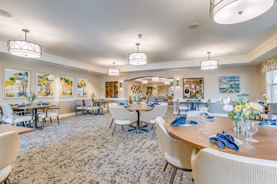 A bright and spacious dining room in an assisted living facility with round and rectangular wooden tables set with blue napkins, glassware, and floral centerpieces. The room features modern light fixtures, patterned carpet, and colorful landscape paintings on the walls.