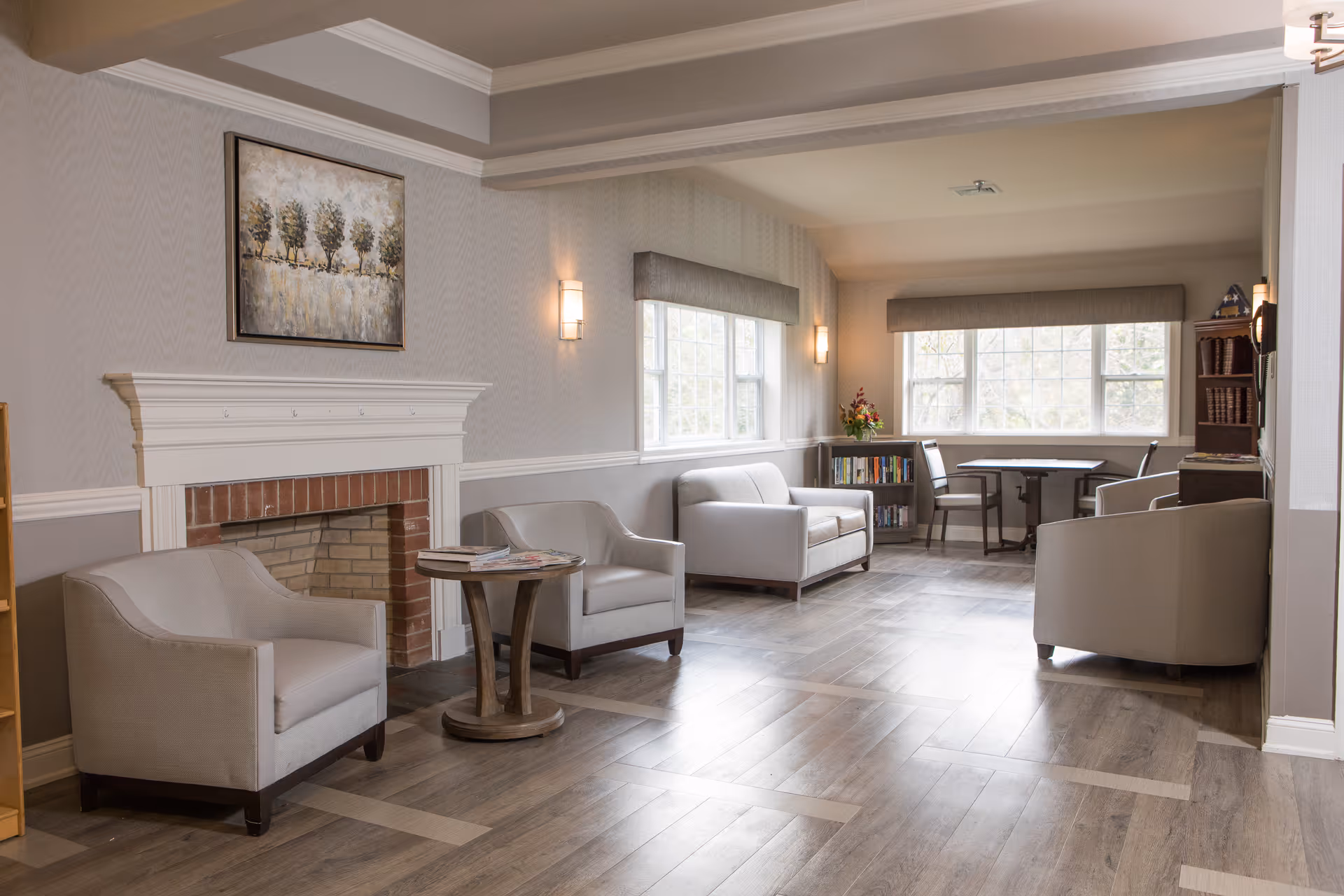 A bright and spacious living room area with light gray armchairs and a loveseat arranged around a small round wooden table. There is a decorative fireplace with a painting of trees above it on the left wall. Large windows with gray valances let in natural light, and a small bookshelf and a table with chairs are visible near the windows. The room has light-colored walls and wood flooring.