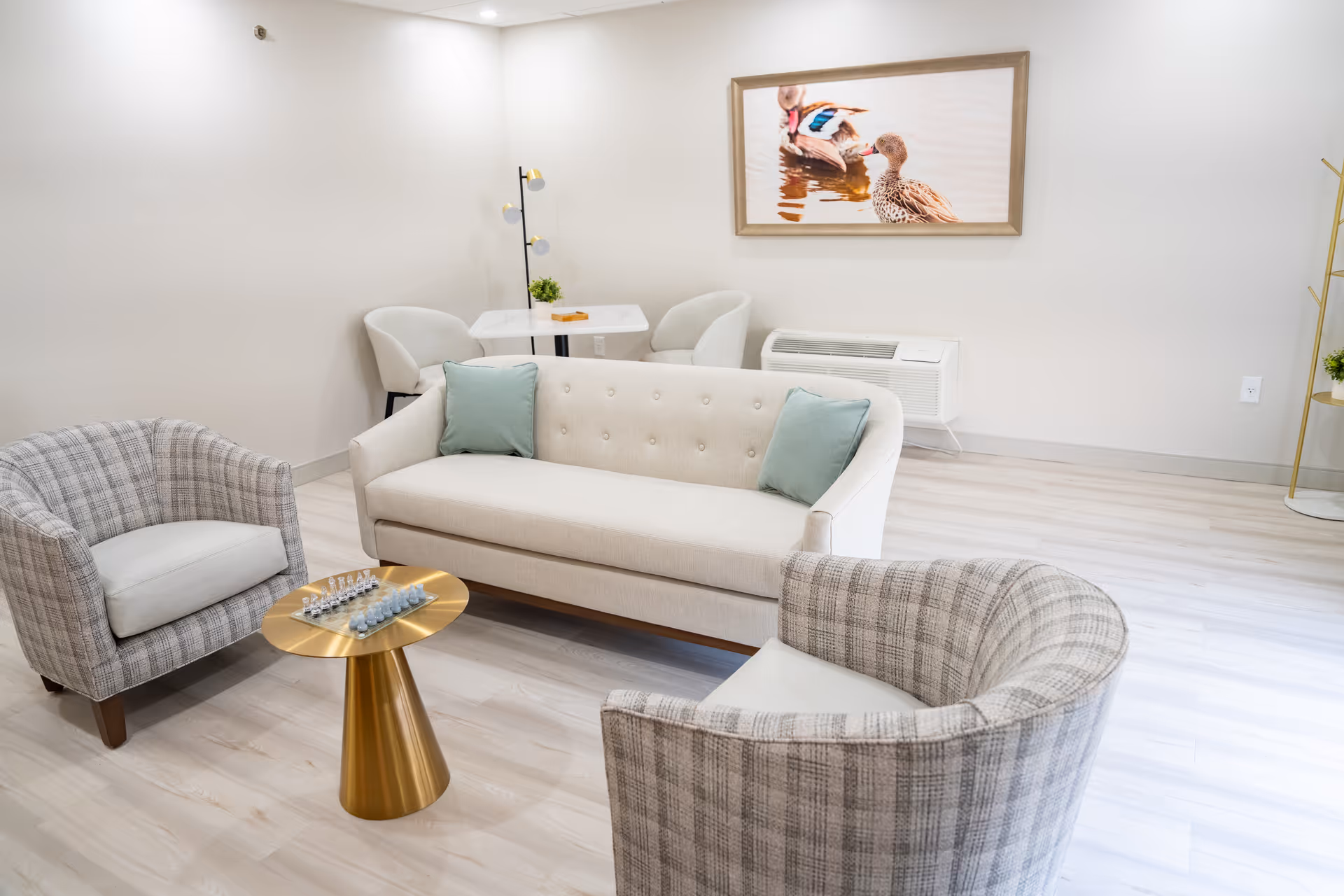 A bright and modern living room area with a beige tufted sofa adorned with two light green pillows, two plaid upholstered armchairs, and a small round gold coffee table with a chess set on it. In the background, there is a small white table with two white chairs, a floor lamp, a wall-mounted air conditioning unit, and a framed picture of ducks on the wall.