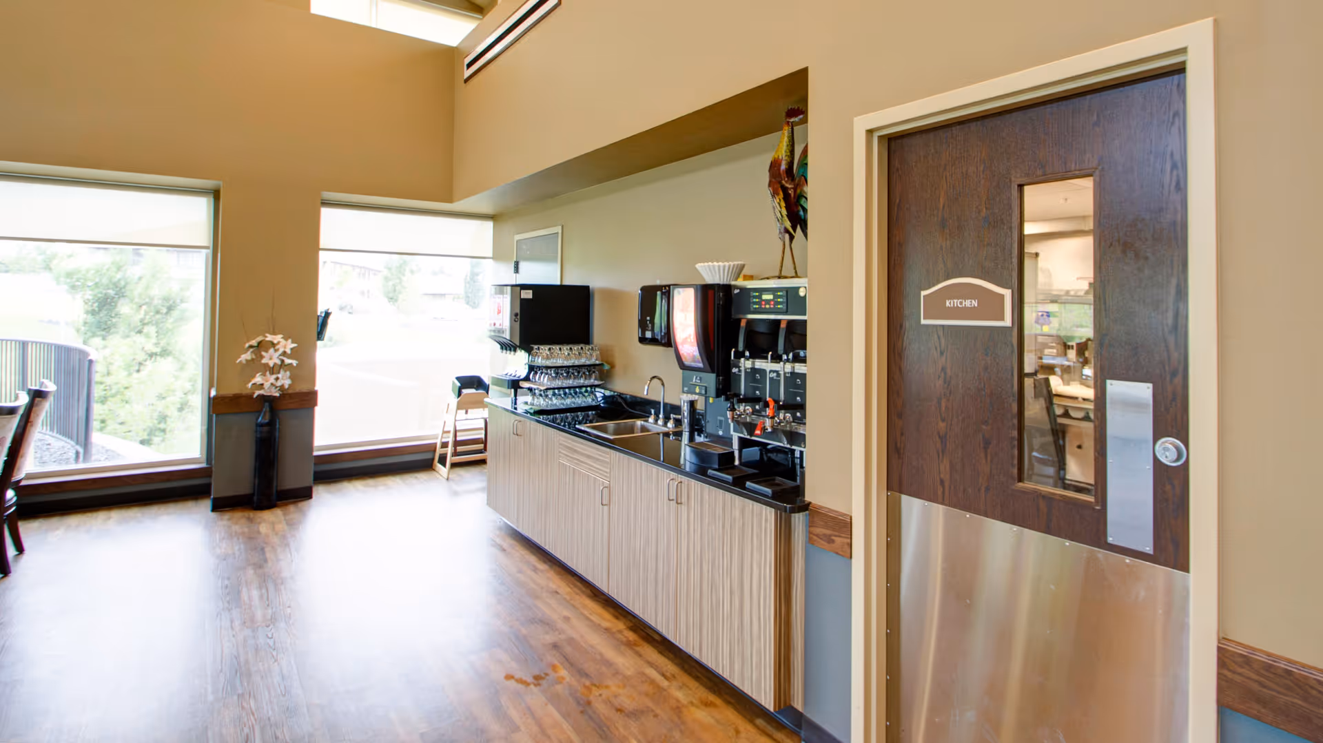 Interior view of a senior living facility area with large windows letting in natural light, a beverage station with coffee and soda machines, a sink, and a door labeled 'KITCHEN'. There is a vase with flowers near the windows and wooden flooring throughout.