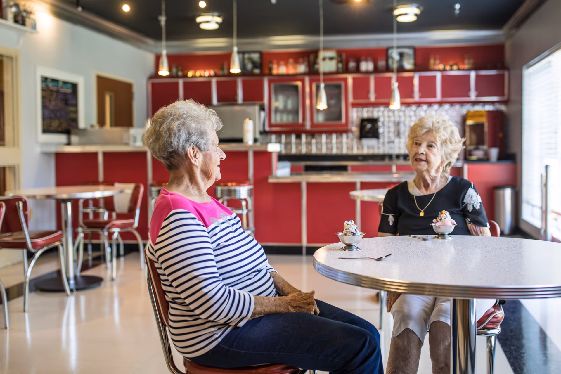 Two elderly women sitting at a round table in a retro-style dining area with red cabinets and pendant lights, each with a small bowl of ice cream topped with sprinkles in front of them.