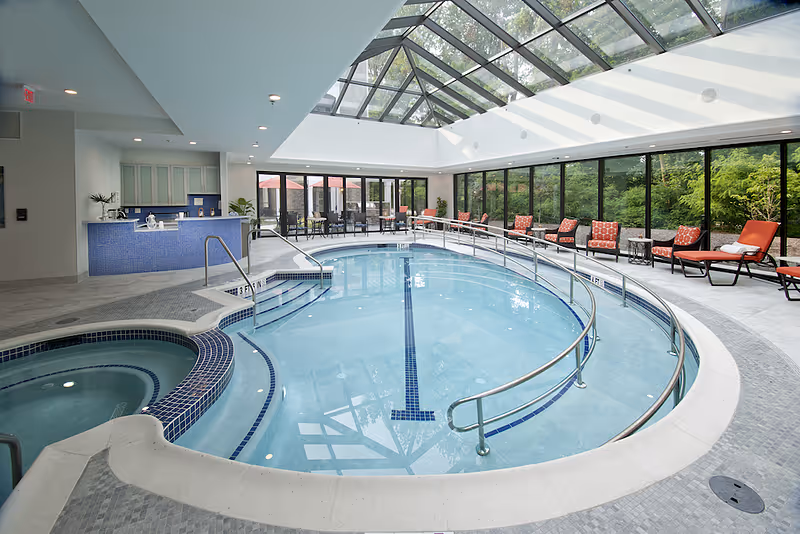 Indoor swimming pool area with a hot tub, surrounded by lounge chairs with red cushions and tables. The space has large glass windows and a glass ceiling allowing natural light to fill the room. There is a blue-tiled counter area in the background.
