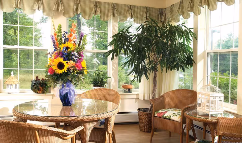 A bright sunroom with large windows showing green trees outside. Inside, there is a round glass-top wicker table with a colorful bouquet of flowers in a blue vase. Surrounding the table are wicker chairs with floral cushions. A tall potted plant is placed near the windows, and decorative items like a small birdhouse and a white lantern are on side tables and window sills.