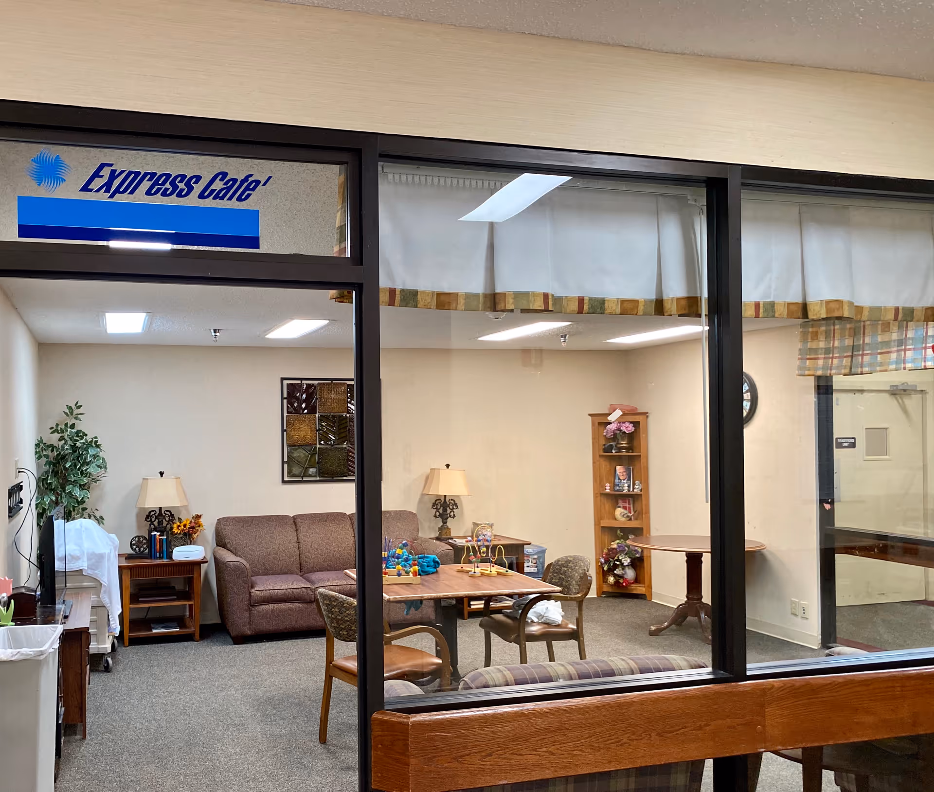View through glass windows into a cozy common area labeled 'Express Cafe' with a brown couch, wooden chairs around a table with toys, a wooden shelving unit with decorative items, two table lamps, and a potted plant in the corner.