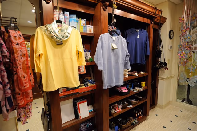 Interior view of a small retail area within a facility, featuring wooden shelves stocked with various items including clothing, personal care products, and small accessories. Three shirts in yellow, light blue, and dark blue hang on display. The floor is tiled, and a glass door with floral decorations is visible on the right side.