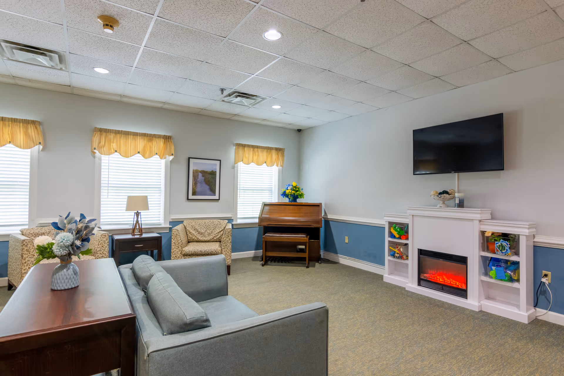 A cozy living room with a gray sofa, two patterned armchairs, a wooden side table with a lamp, a piano with a flower vase on top, a wall-mounted flat screen TV above a white electric fireplace, and built-in shelves with colorful toys. The room has three windows with yellow valances and a framed picture on the wall.