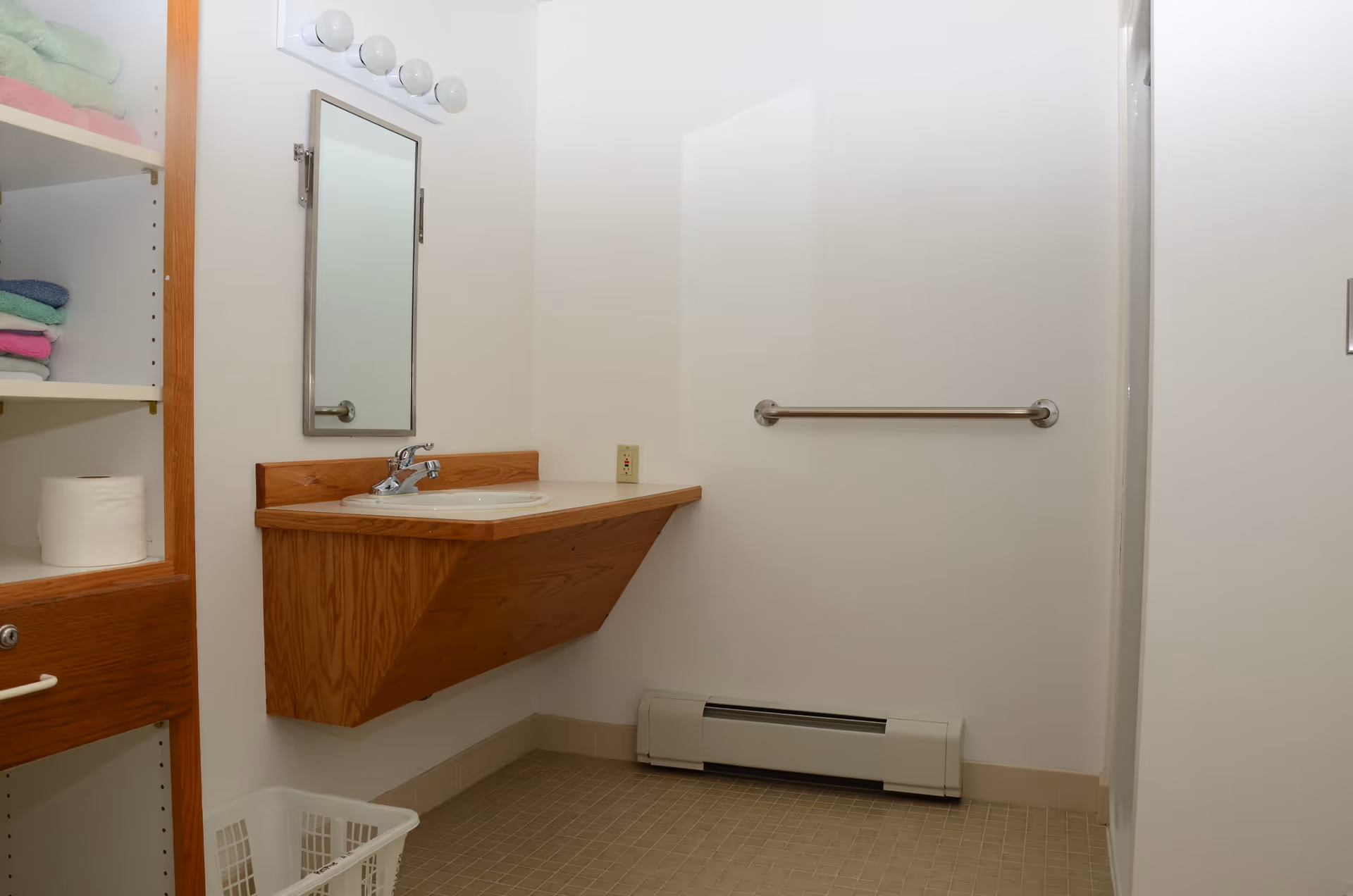 A small bathroom with a wall-mounted wooden sink and faucet, a rectangular mirror above the sink with four round light bulbs above it, a metal grab bar on the wall, a white baseboard heater below the grab bar, and open shelving on the left side holding folded towels and a roll of toilet paper. There is a white laundry basket on the floor.