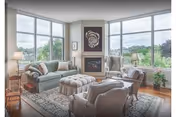 A bright and spacious living room with large windows offering a scenic outdoor view. The room features a green sofa, two armchairs, a cushioned ottoman, a patterned area rug, a fireplace with artwork above it, and two table lamps on either side of the sofa. There are wooden floors and a few decorative plants near the windows.