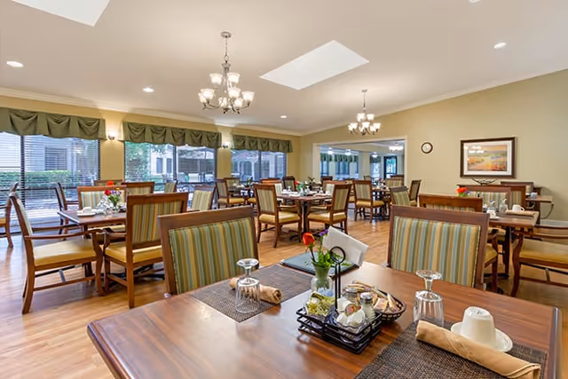 Spacious senior living dining room with multiple wooden tables and striped-upholstered chairs set for meals, large windows, chandeliers, and wood flooring.