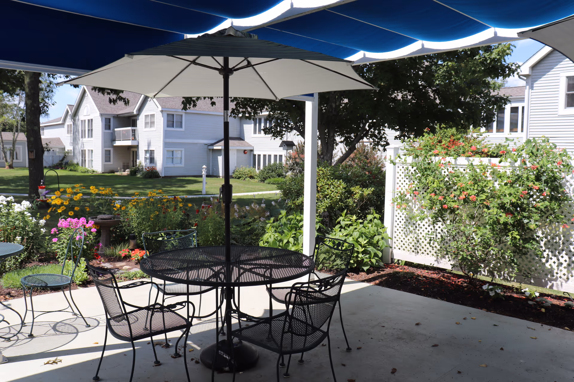 Outdoor patio area with a round metal table and four matching chairs under a large white umbrella. The patio is surrounded by colorful flower beds and greenery, with residential buildings visible in the background under a clear sky.