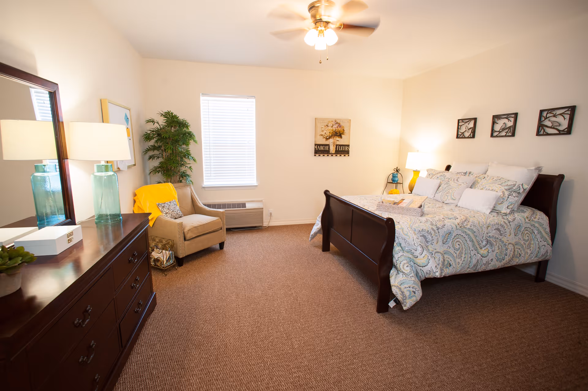 A cozy bedroom with a wooden bed frame and a patterned comforter, a beige armchair with a yellow throw blanket, a wooden dresser with a large mirror, two blue glass lamps, a potted plant, and wall decorations including a floral painting and three framed bird silhouettes.
