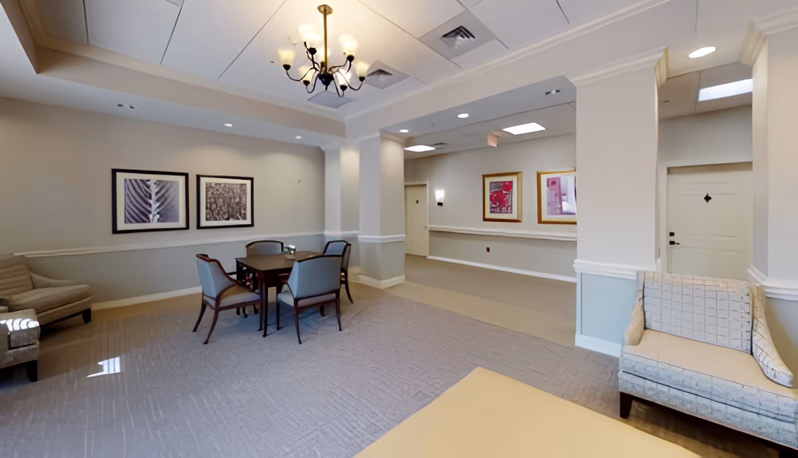 Bright common area with a table and chairs, lounge seating, framed artwork, and a chandelier in a senior living facility.