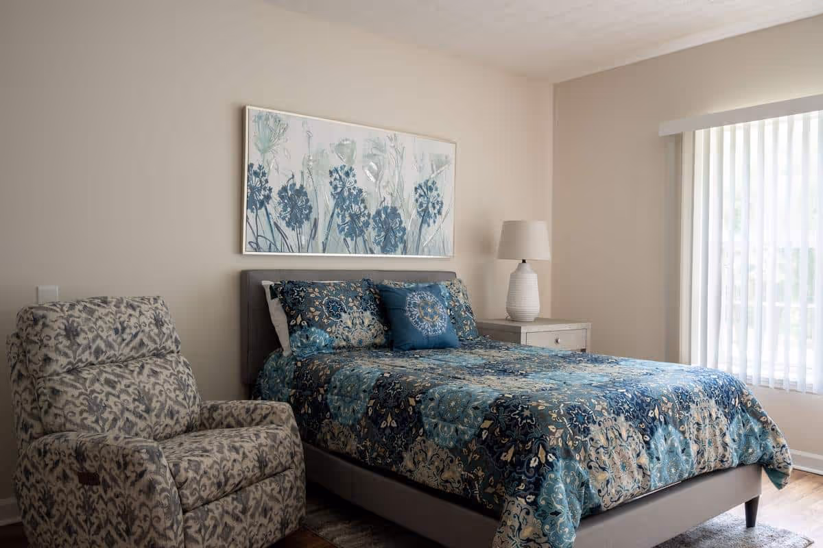 A bedroom with a bed covered in a blue and beige floral patterned comforter and matching pillows. To the left of the bed is a patterned armchair, and to the right is a nightstand with a white lamp. A large window with vertical blinds is on the right wall, and a framed floral artwork hangs above the bed.