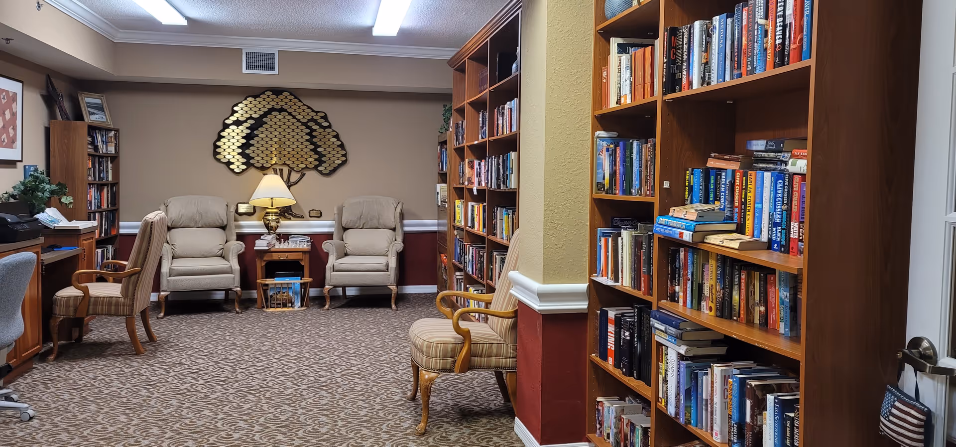 A cozy library or reading room with multiple wooden bookshelves filled with books. There are several upholstered armchairs and a small table with a lamp in the center against the back wall. The room has patterned carpet and warm lighting, creating a comfortable atmosphere.