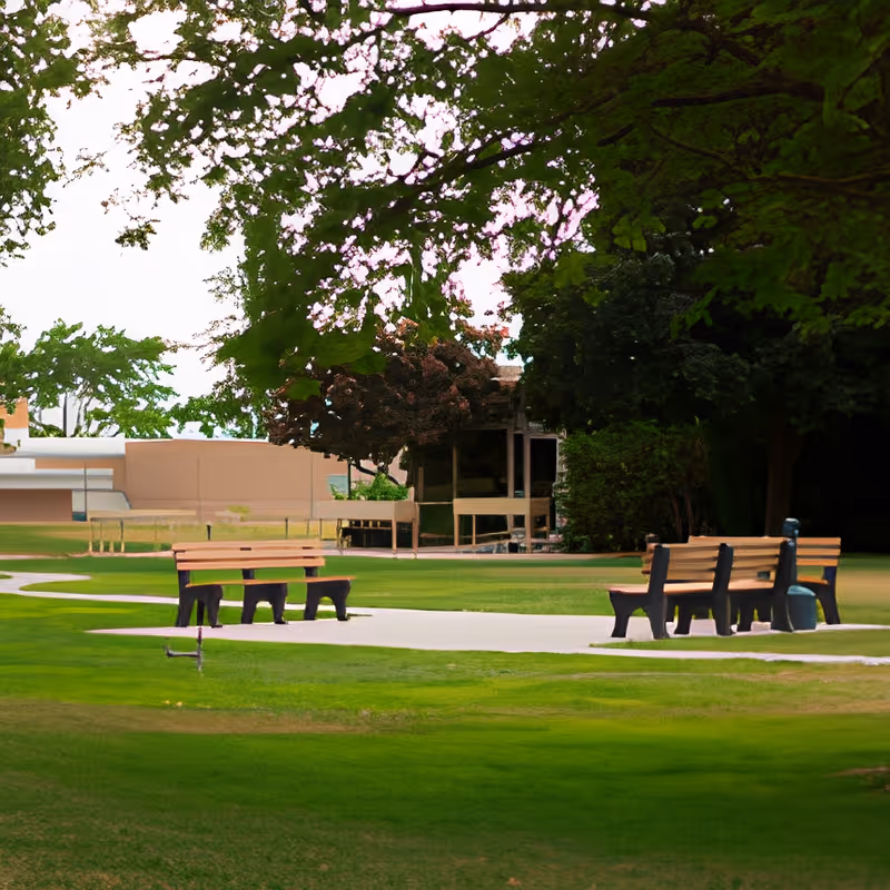 Park-like outdoor area with benches along paved paths, green lawn and large trees with buildings in the background.