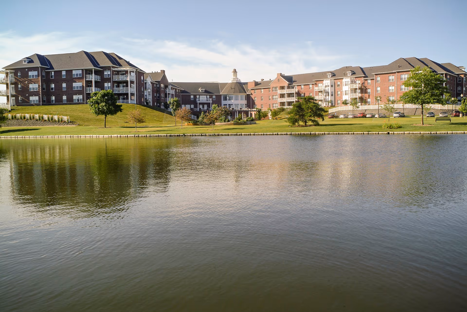 View of a large senior living facility named Immanuel: The Landing, featuring multiple connected brick buildings with balconies, situated on a grassy hill beside a calm body of water under a partly cloudy sky.