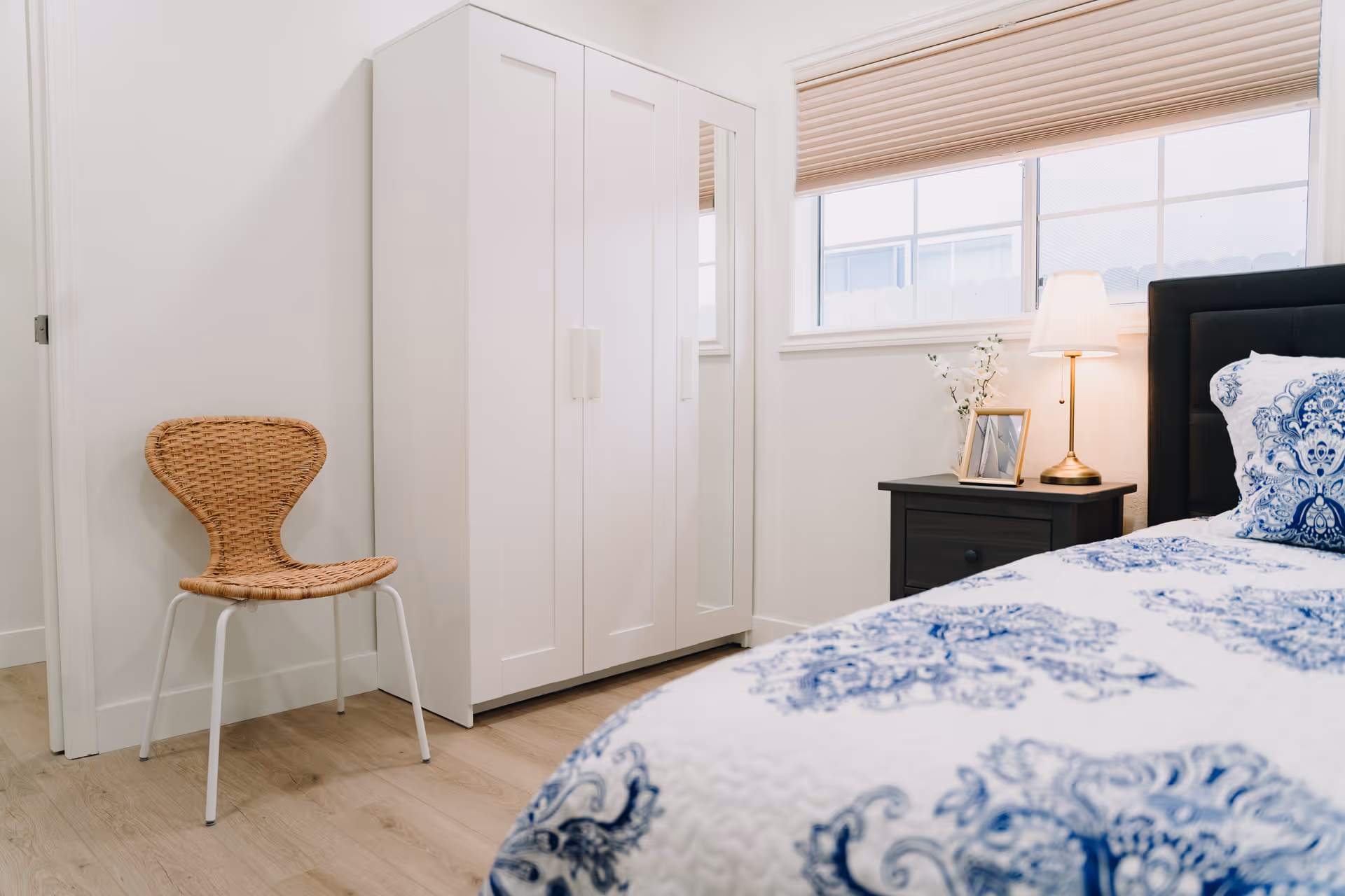 Bright bedroom with blue-patterned bedding, a bedside table and lamp, a white wardrobe, and a wicker chair.