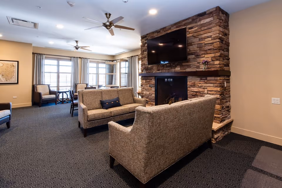 A cozy senior living common area with two beige sofas facing a stone fireplace with a mounted flat-screen TV above it. The room has large windows with curtains, two ceiling fans, and additional seating including armchairs and a small table in the background.