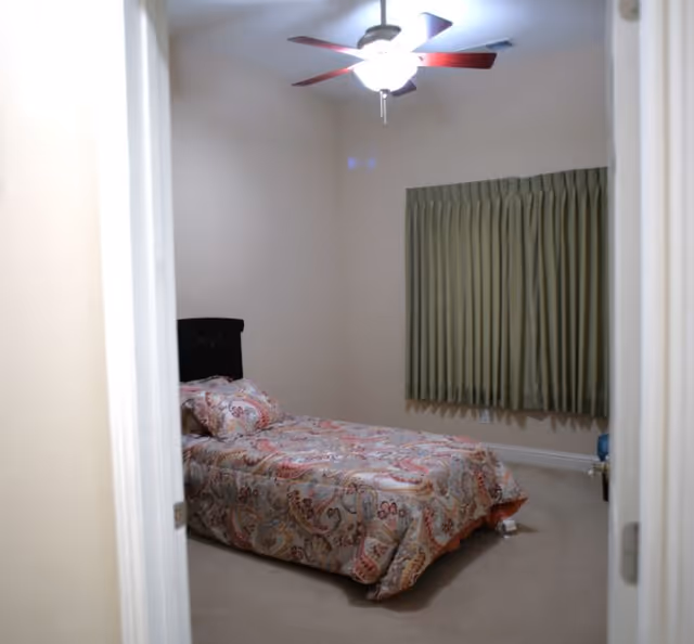 Small bedroom with a single bed covered in a patterned comforter, green curtains, and a ceiling fan overhead.