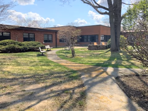 Outdoor courtyard area of a brick building with a curved concrete pathway, benches, bushes, trees, and a clear blue sky with some clouds.