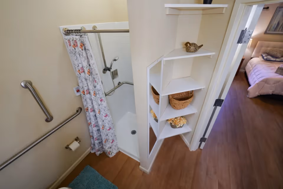 A small bathroom with a shower area featuring a floral shower curtain and grab bars on the walls. Adjacent to the bathroom is a corner shelving unit with decorative items and baskets. The image also shows a glimpse of a bedroom with a bed and pillows through an open door.