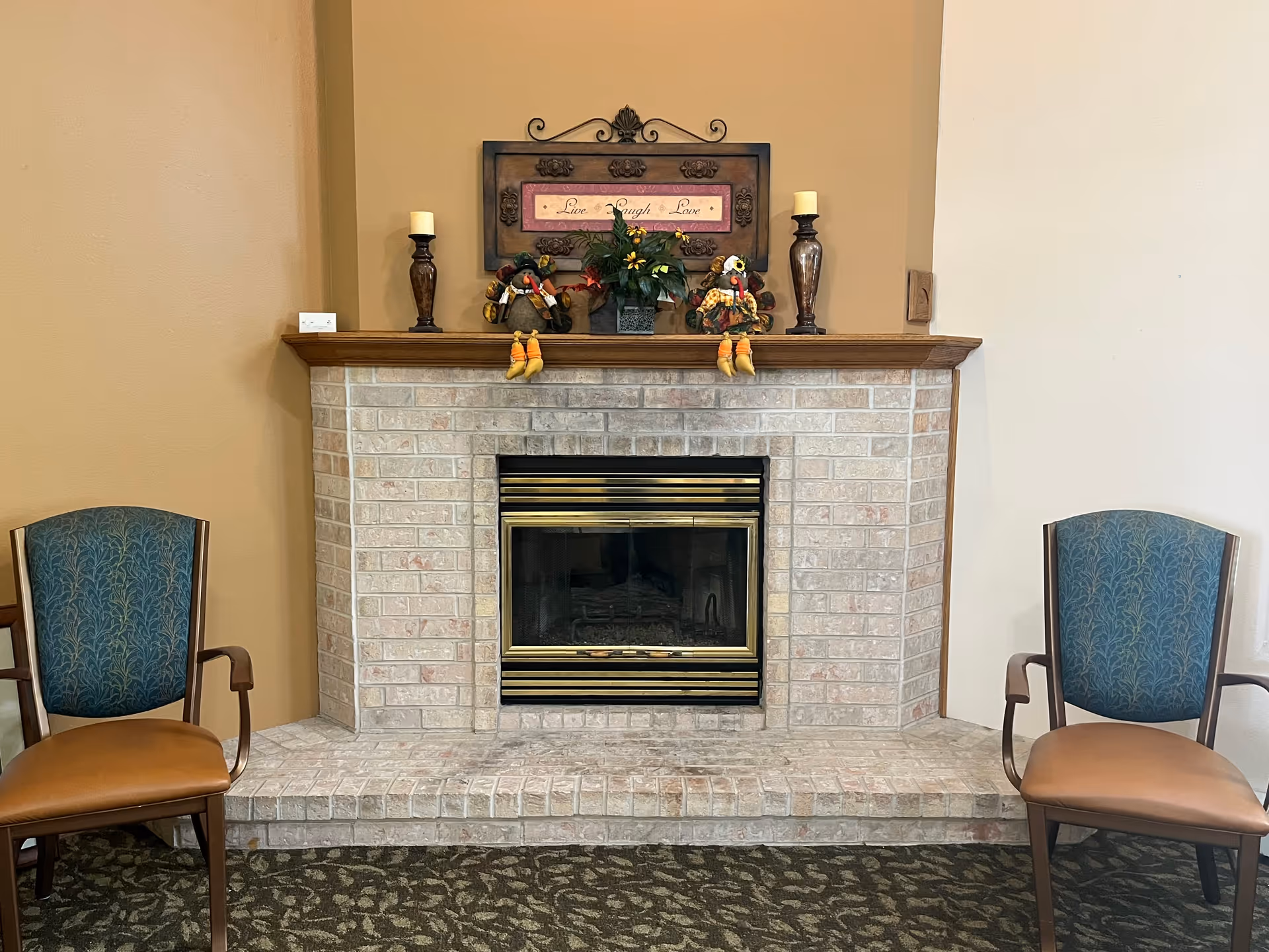 A cozy indoor fireplace with a brick surround and a wooden mantel. On the mantel, there are two decorative candles, two small turkey figurines, a potted plant, and a framed sign that reads 'Live Laugh Love'. Two upholstered chairs with wooden arms and legs are positioned on either side of the fireplace. The walls are painted in warm beige tones and the floor is covered with a patterned carpet.