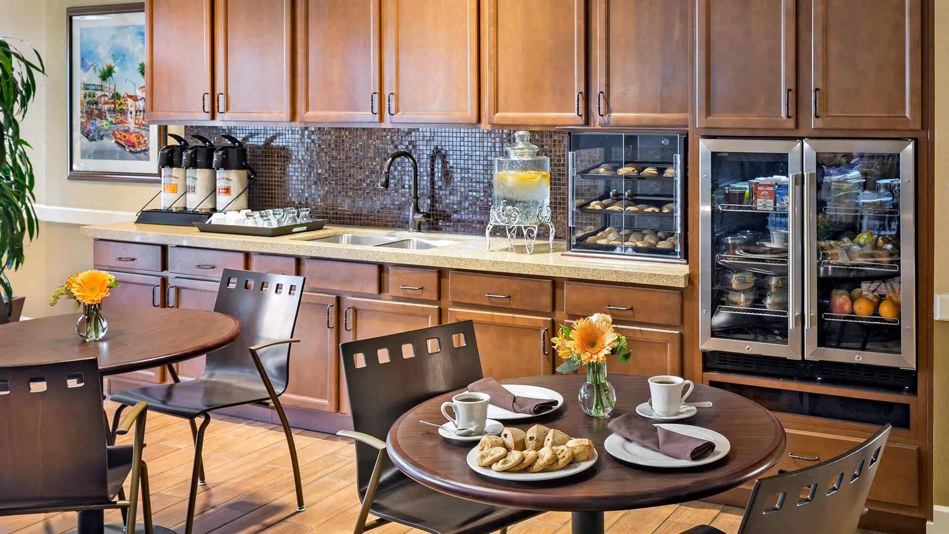 A cozy kitchen area with wooden cabinets and a countertop featuring a water dispenser with lemon slices, a small oven with baked goods, and a glass-front refrigerator stocked with food items. In the foreground, there are two round tables with dark chairs, each table set with a small vase of yellow flowers, plates of cookies, cups, and napkins.