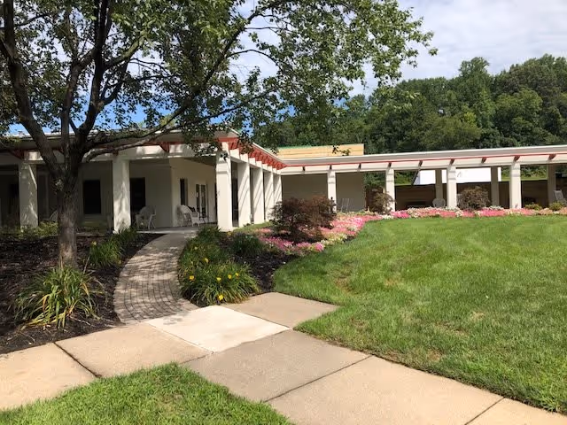 Outdoor view of Regency Park Senior Living Community showing a curved walkway leading to a covered patio area with white columns. The area is landscaped with green grass, flowering plants, and trees under a partly cloudy sky.