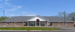 Front exterior view of a single-story brick building with a gray roof, a central entrance with a small portico, and a circular window above the door. The building is surrounded by a grassy area and a paved road in front under a clear blue sky.