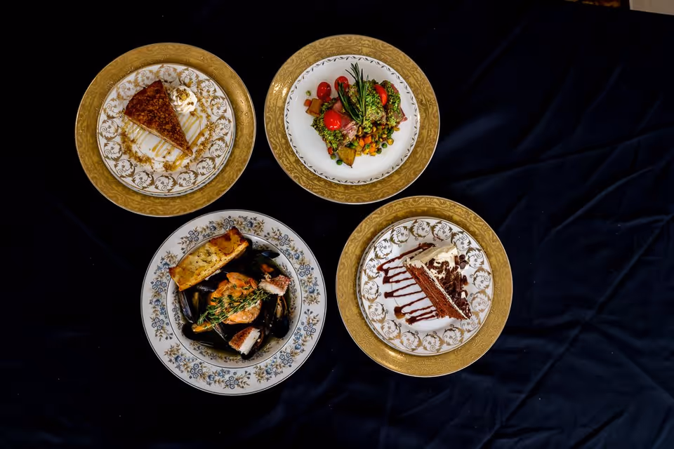 Four plates of food arranged on a dark cloth background. Two plates have slices of cake with decorative drizzle and whipped cream, one plate has a fresh salad with cherry tomatoes and herbs, and one plate contains a seafood dish with bread and garnished with herbs.