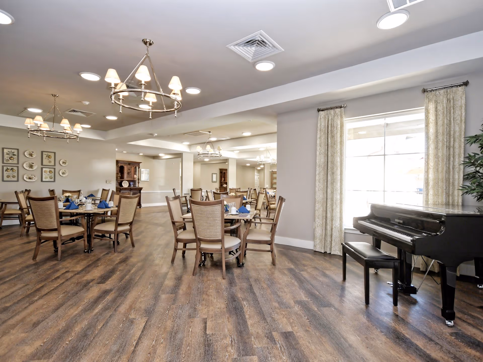 A spacious senior living dining room with multiple round tables set with blue napkins and white dishes. The room features wooden flooring, beige walls, and large windows with patterned curtains allowing natural light. There are elegant chandeliers hanging from the ceiling and a black grand piano with a bench near the window.