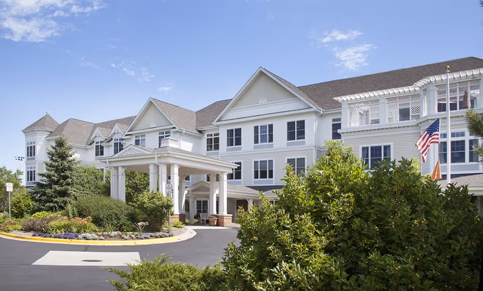Exterior view of a large, white senior living facility building with multiple windows and a covered entrance. The building is surrounded by well-maintained landscaping including bushes, trees, and flowers. An American flag is visible on a flagpole near the entrance under a clear blue sky.