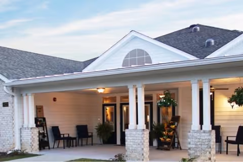 Covered front entrance of a single-story senior living building with columns, seating, potted plants, and hanging baskets.
