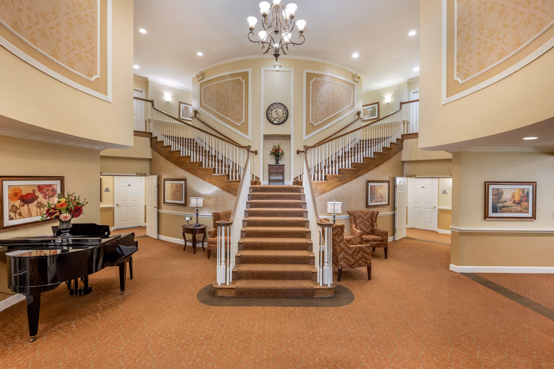 Spacious and elegant interior of a senior living facility featuring a central carpeted staircase with two symmetrical side staircases, a chandelier hanging from the ceiling, a black grand piano on the left, two armchairs with side tables and lamps on either side of the main staircase, and framed artwork on the walls.