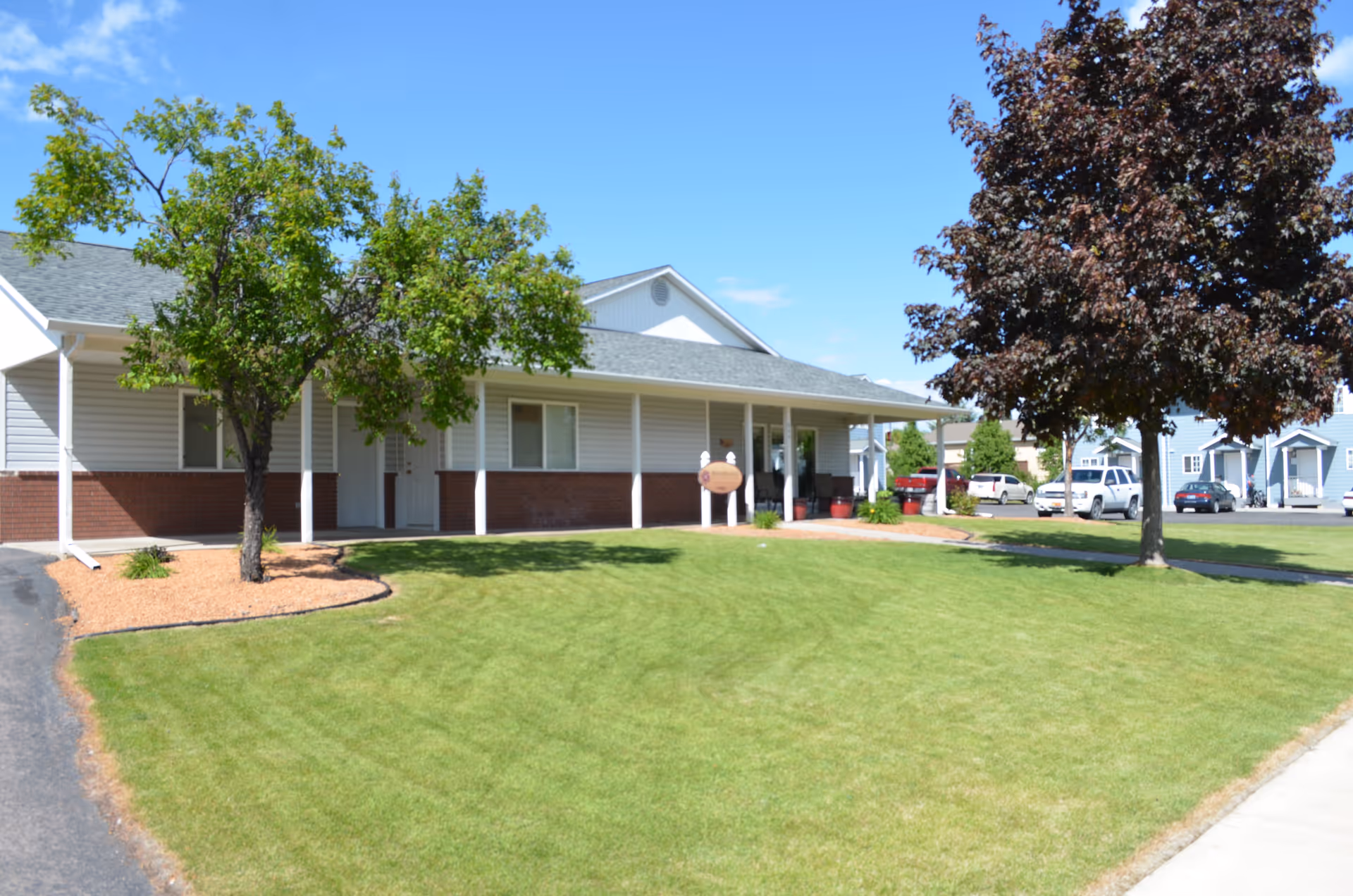 Single-story building with white siding and brick accents, a covered porch, and two trees in the front yard with green grass under a clear blue sky. Several parked cars and neighboring houses are visible in the background.