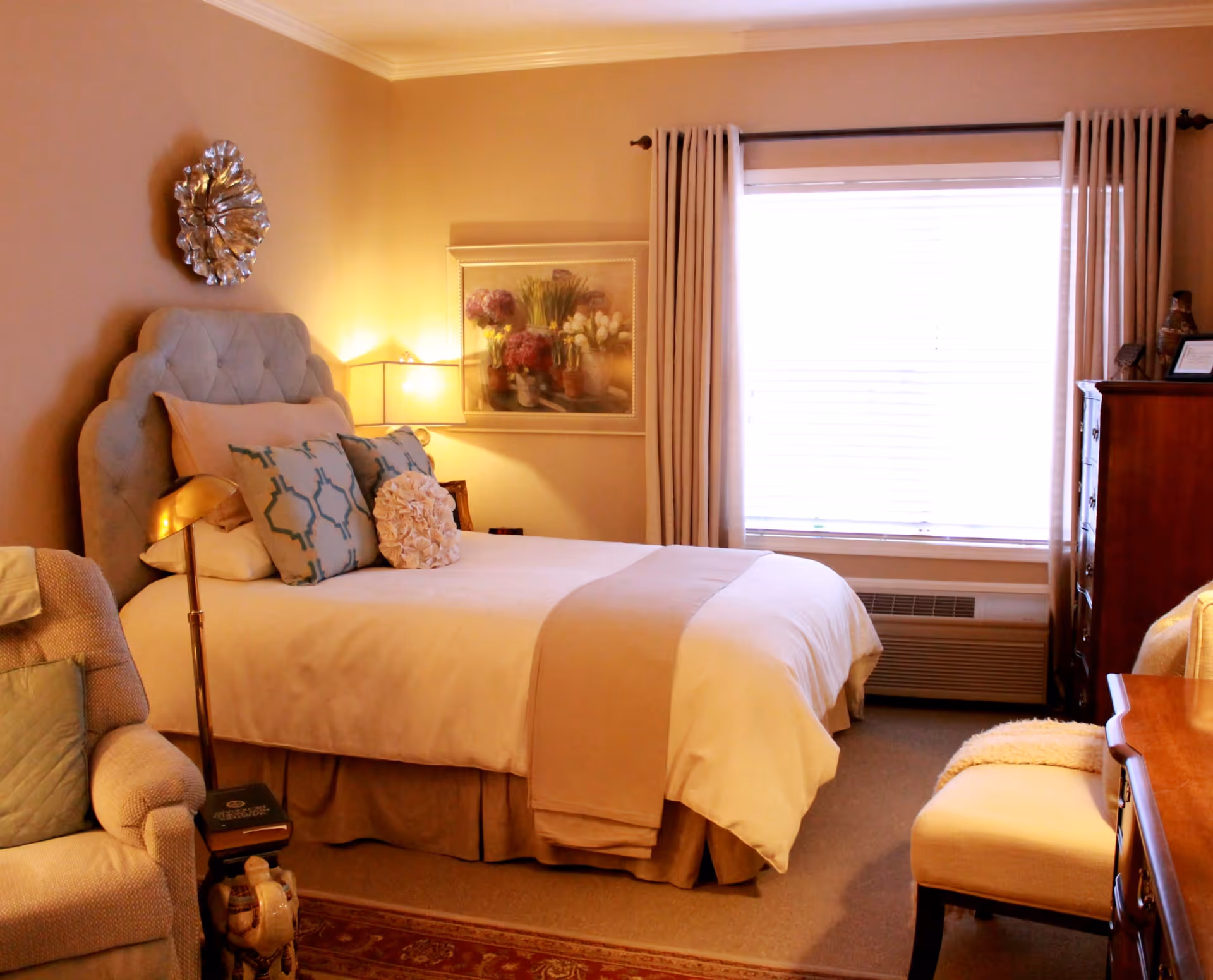 A cozy bedroom in an assisted living facility featuring a neatly made bed with multiple decorative pillows and a beige throw blanket. The room includes a cushioned armchair with a pillow, a wooden dresser, a window with closed blinds and beige curtains, a bedside lamp, and wall art depicting flowers. The room is warmly lit and decorated in soft neutral tones.