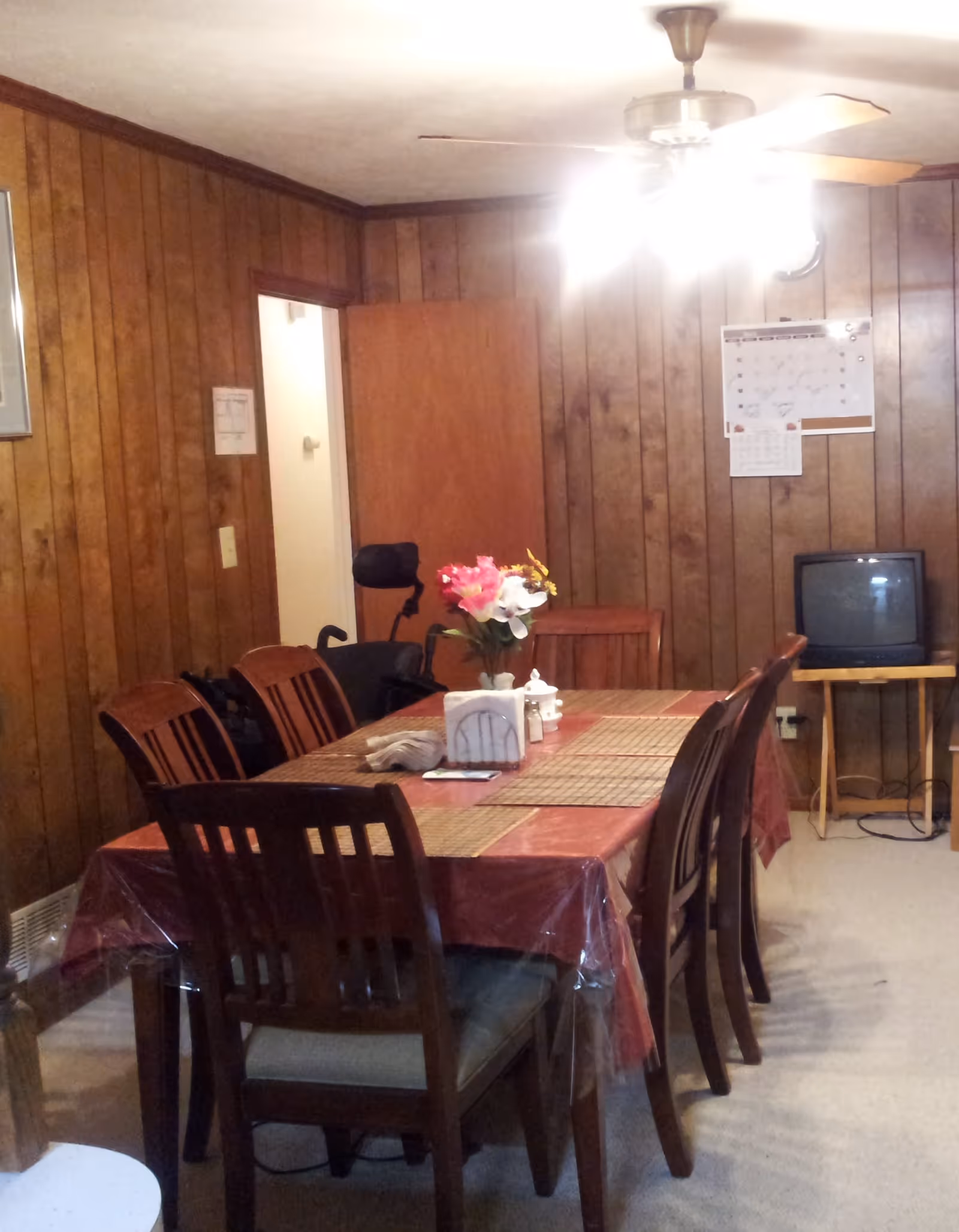 A dining room with wood-paneled walls and a ceiling fan with lights. There is a rectangular dining table covered with a red tablecloth and a clear plastic cover, set with placemats, a vase with flowers, napkins, and condiments. Six wooden chairs surround the table. In the background, there is an open door, a wheelchair, a small TV on a wooden stand, and a calendar on the wall.
