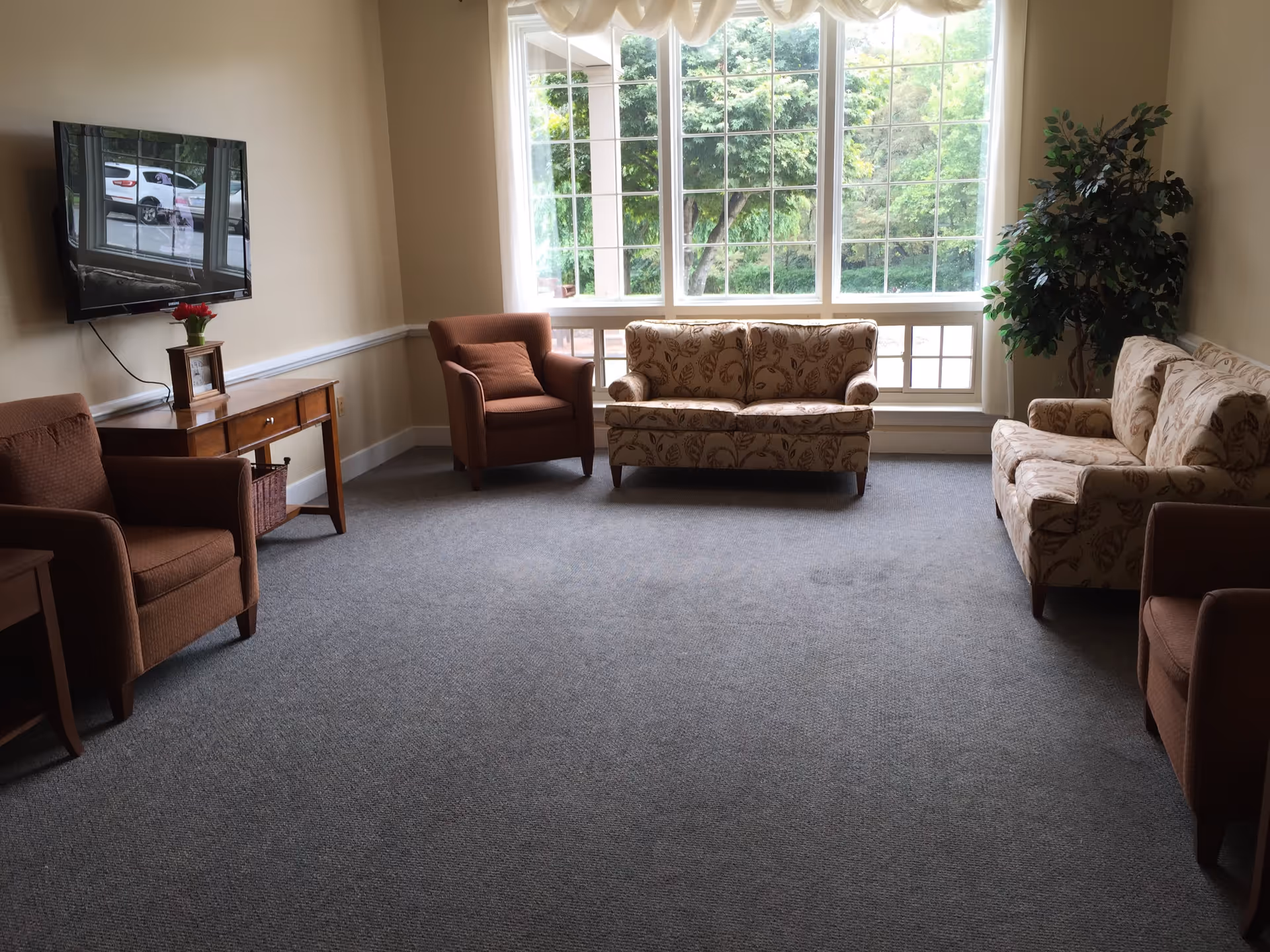 Bright common living room with sofas and armchairs arranged around a large window and a wall-mounted TV.